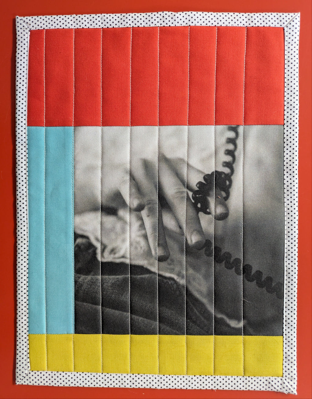 Artistic quilt with a central black and white photo of a hand holding a phone, surrounded by colorful borders on a red background.