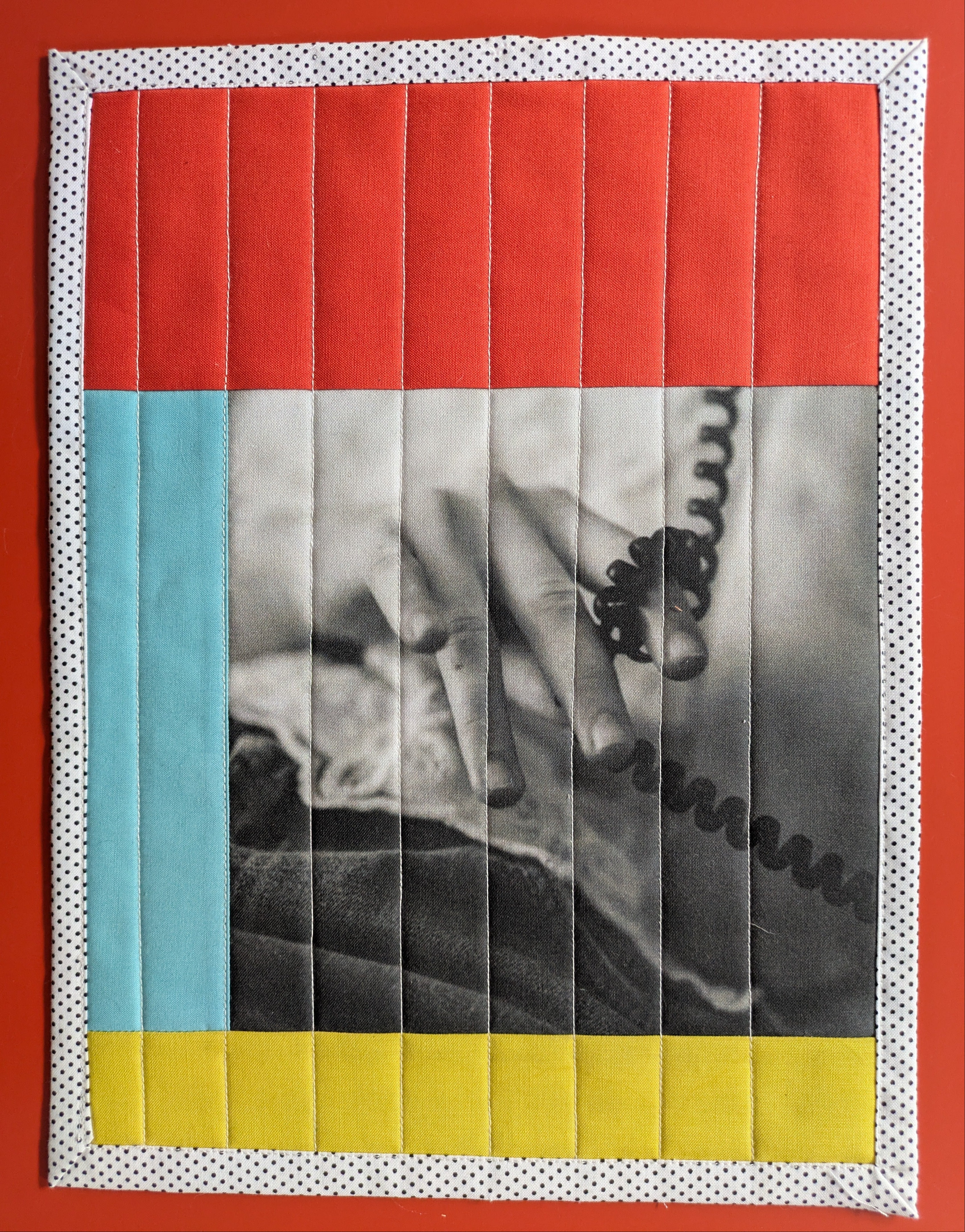 Artistic quilt with a central black and white photo of a hand holding a phone, surrounded by colorful borders on a red background.
